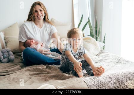 Mama mit Kindern auf dem Bett. Das Baby ist in den Armen der Mutter, das Kleinkind sitzt neben ihm, beleidigt. Zwei Kinder in einer Familie, die Eifersucht der Kinder auf thei Stockfoto