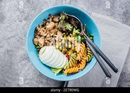 Gesunde Schüssel zum Mittagessen. Hühnchen, Nudeln Fusilli, mischen Grüns, grüne Erbsen und Sonnenblumenkerne in blauer Platte auf Beton grauen Hintergrund Tisch. Köstlicher Bala Stockfoto