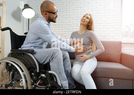 Behinderter afroamerikanischer Mann, der den Bauch seiner Schwangeren berührt kaukasische Frau, während sie auf dem Sofa zu Hause entspannen Stockfoto