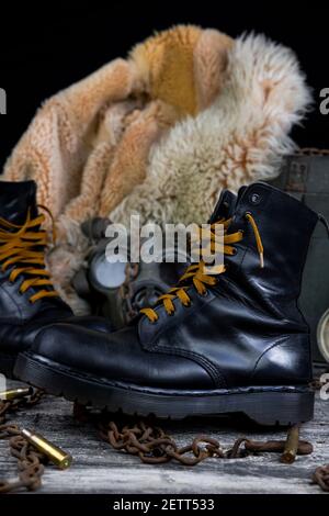 Lederstiefel mit verrosteten Ketten und Schussschalen mit Militär Gasmaske und Pelzmantel im Hintergrund ruhen auf Holz Tabelle Stockfoto