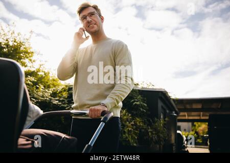 Vater schiebt auf Baby Kinderwagen reden auf Handy im Freien. Mann auf Vaterschaftsurlaub mit seinem Baby im Kinderwagen und im Gespräch auf Handy. Stockfoto