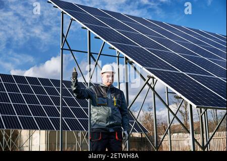 Männlicher Techniker zeigt Genehmigung Geste Daumen nach oben, während in der Nähe von Solar-Photovoltaik-Panel-System stehen. Elektriker genehmigt alternative Energiequellen. Konzept der Macht nachhaltige Ressourcen. Stockfoto