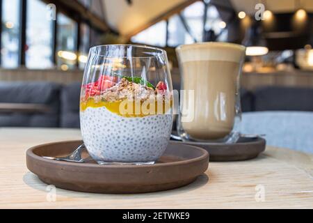 Chia Pudding mit Mangopüree, Haferflocken und frischen Erdbeeren und Tasse mit Cappuccino auf Holztisch im Café Selective Focus. Stockfoto