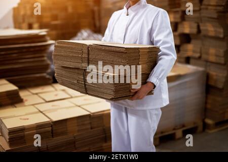 Nahaufnahme eines jungen, hart arbeitenden Mitarbeiters in sterilen Tüchern, der den Stapel aus braunen Faltschachteln aus Pappe aus dem Laderaum der Fabrik transportiert. Stockfoto