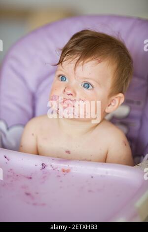 Adorable Baby essen auf Babystuhl und machen ein Durcheinander. Kleine Mädchen essen Nahrung. Nettes Baby mit einem lustigen Ausdruck auf seinem Gesicht. Stockfoto