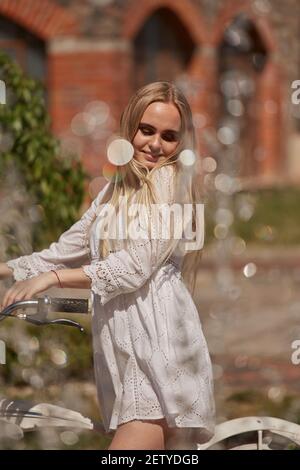 Hübsche blonde Frau in weißem Kleid mit Fahrrad in der Nähe Brunnen Bei sonnigem Licht Stockfoto