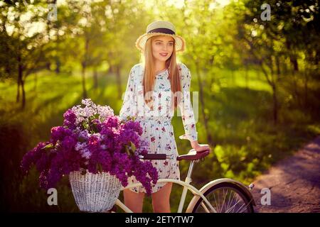 Porträt einer glücklichen schönen Frau in Strohhut mit Vintage-Fahrrad und Blumen auf Park Hintergrund im Sonnenlicht im Freien. Fahrrad mit Korb voller Blumen. Active Leisure-Konzept. Stockfoto