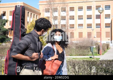 Lateinamerikanische Freunde tragen eine schützende Gesichtsmaske und gehen mit einem Gitarrenkoffer um den Universitätscampus. Neue Normalität auf der Straße. Stockfoto