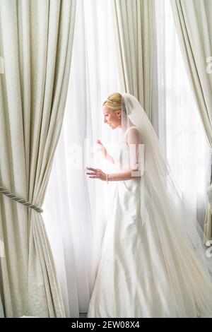 Braut, die aus dem Fenster durch die Voile Vorhänge schaut. Sie schaut auf alle Gäste, die ankommen und lächeln. Stockfoto