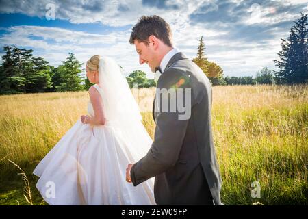 Braut und Bräutigam frisch vermählte, die durch das lange Gras des Herrenhauses Gelände gehen, kurz nachdem sie geheiratet haben.die Braut ist führt den Spaziergang. Stockfoto