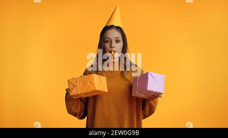 Teenager-Mädchen in Party Cap Blick auf Kamera beim Blasen Party-Horn und halten Geschenke isoliert auf gelb Stockfoto