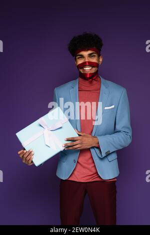 Happy african american Mann gebunden mit roten Band halten präsent Auf violett Stockfoto