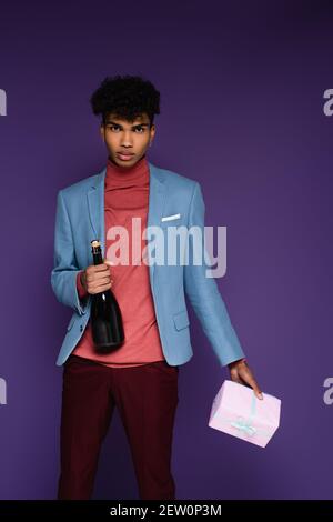 Junger afroamerikanischer Mann in blauem Blazer hält verpackt Geschenk Und eine Flasche Champagner auf Purpur Stockfoto