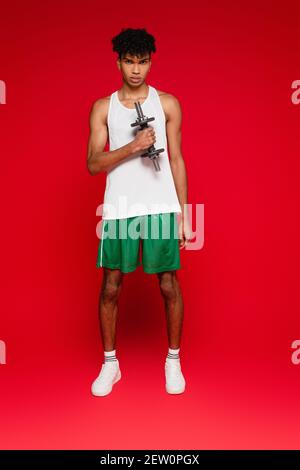 Volle Länge des sportlichen afroamerikanischen Mannes in Shorts und Tank Top Training mit Hantel auf rot Stockfoto