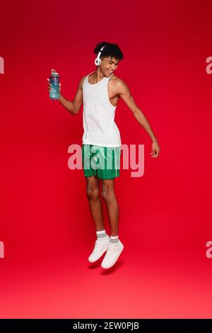 In voller Länge lächelnder afroamerikanischer Mann in kabellosen Kopfhörern Halten Sportflasche beim Springen auf rot Stockfoto