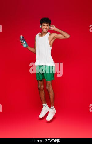In voller Länge glücklicher afroamerikanischer Mann in kabellosen Kopfhörern Halten Sportflasche beim Springen auf rot Stockfoto