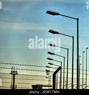 Silhouette von Straßenlaternen vor dem Metallzaun mit Stacheldraht mit Licht Schleier Wolken in blauen Himmel in Hintergrund Stockfoto