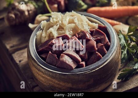 Hoher Winkel der frischen rohen Hähnchenleber mit Knorpeln geschnitten In hölzernen PET Schüssel auf Tisch mit ungekochten reifen platziert Gemüse Stockfoto