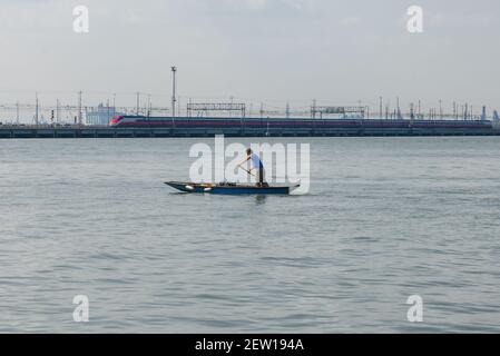 VENEDIG, ITALIEN - 27. SEPTEMBER 2017: Ein Mann ruht auf einem alten Holzboot vor dem Hintergrund eines Hochgeschwindigkeitszuges. Lagune Von Venedig Stockfoto