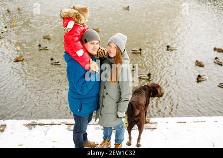 In voller Höhe Familienportrait mit einem Kind und Hund im Winter Casual Outfit posiert im Freien in der Nähe des Flusses mit Entenvögeln, Wochenende Aktivitäten und frische Luft Stockfoto