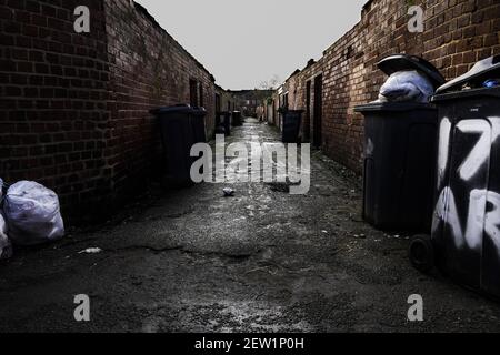 Eine Seitenstraße oder Gasse zwischen heruntergekommenen Reihenhäusern Im Norden Englands mit Mülltonnen und Wheelie Behälter warten auf Abholung Stockfoto