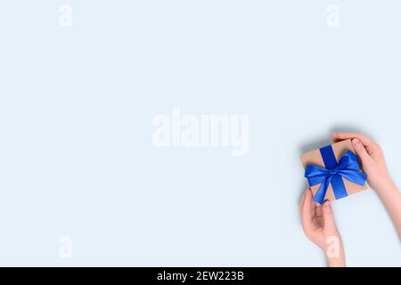Weibliche Hände halten verpackte Geschenkbox auf einem blauen pastellfarbenen Hintergrund. Geschenk gebunden mit einem Band. Stockfoto