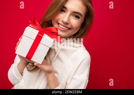 Nahaufnahme Foto Aufnahme von attraktiven glücklich lächelnd blonde junge Frau Isoliert über bunten Hintergrund Wand trägt weißen Hoodie mit weißem Geschenkbox Stockfoto