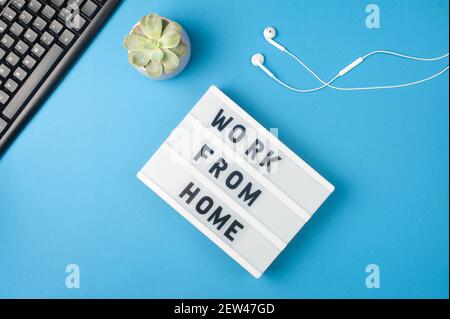 Arbeiten von zu Hause aus - Text auf dem Leuchtkasten mit blauem Hintergrund am Arbeitsplatz. Schwarze Tastatur und weiße Kopfhörer. Konzept für freiberufliche Arbeit Stockfoto