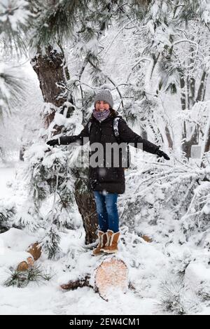 Lächelnde junge Frau, die auf einem umgestürzten Baum nach Schneelast und Schnee in einem schneebedeckten Winterpark steht. Mädchen genießen verschneiten Winter, frostigen Tag. Gehen Sie hinein Stockfoto