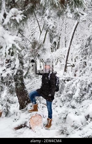 Lächelnde junge Frau, die auf einem umgestürzten Baum nach Schneelast und Schnee in einem schneebedeckten Winterpark steht. Mädchen genießen verschneiten Winter, frostigen Tag. Gehen Sie hinein Stockfoto