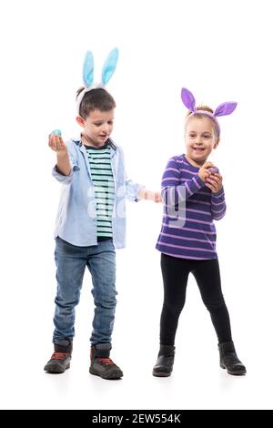 Nette verspielte kleine Junge und Kleinkind Mädchen mit Osterhasen Ohren spielen Ei klopfen Spiel. Ganzkörper isoliert auf weißem Hintergrund. Stockfoto