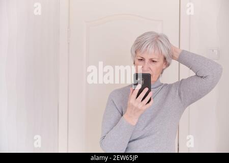 Eine grauhaarige ältere Frau in einem grauen Rollkragen, die zu Hause aufmerksam auf das Smartphone schaut. Lifestyle-Moment. Stockfoto