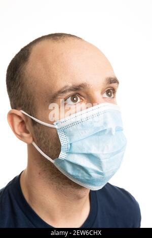 Ein kaukasischer Mann trägt ein blaues T-Shirt und eine chirurgische Gesichtsmaske mit einem neutralen Ausdruck, der nach links schaut. Weißer Hintergrund. Stockfoto