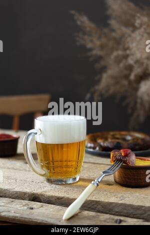 Glas mit Bier und Schaum und Gabel mit einem Stück Von Wurst und Ketchup auf Holzgrund, deutsche Bratwürste In einem Teller und Ketchup mit Senf Stockfoto