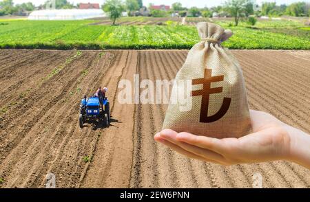 Hand mit einer türkischen Lira Geldbeutel auf dem Hintergrund eines Bauernfeldes mit einem Traktor. Beihilfen für landwirtschaftliche Erzeuger. Grundbesitz, Land ma Stockfoto