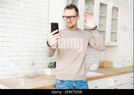 Portrait von charmanten jungen Mann zu Hause stehen, Begrüßung durch Videoanruf, im Gespräch auf dem Handy, mit einem Team-Meeting, Aufnahme von Video, ein Selfie-Foto, Freiberufler, aufgerufen, Guten Morgen zu sagen Stockfoto