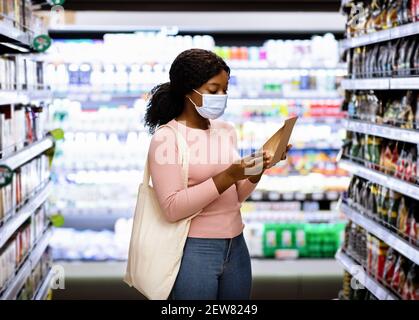 Millennial schwarze Dame mit Tragetasche trägt Gesichtsmaske, Einkaufen für Lebensmittel im Einkaufszentrum während covid Ausbruch Stockfoto