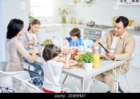 Fröhliche hispanische Familie, die zusammen essen, während sie ein Frühstück genießen, am Tisch in der Küche zu Hause sitzen. Kindheit, Essen Konzept Stockfoto