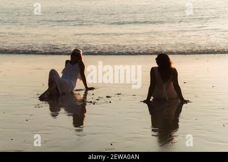 Junge Freundinnen in weißen Kleidern sitzen am Strand Stockfoto