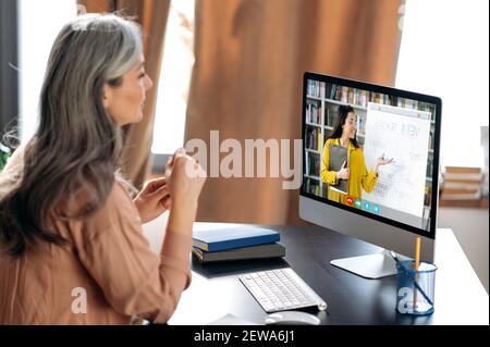 Online-Schulungen. Grauhaarige Frau mittleren Alters, die online per Videoanruf studiert, benutzt App und Computer, sitzt an ihrem Schreibtisch, hört sich eine Online-Lektion an, auf dem Bildschirm der Lehrerin in der Nähe des Stockfoto