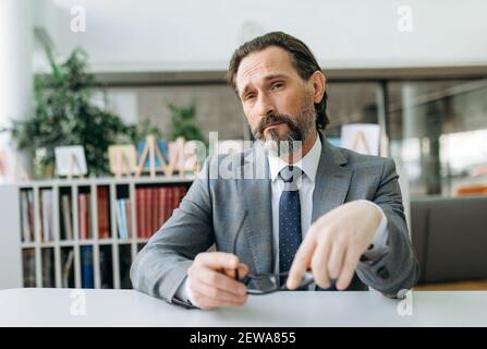 Porträt von gutaussehenden mittleren Alters Geschäftsmann in stilvollen eleganten Anzug am Arbeitsplatz. Einflussreicher erfolgreicher Unternehmer sitzt im modernen Büro und blickt direkt auf die Kamera Stockfoto