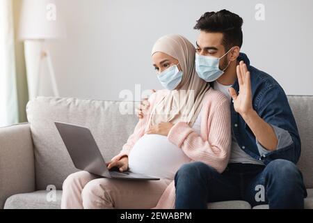 Schwangere muslimische Paar trägt medizinische Masken machen Videoanruf mit Laptop zu Hause Stockfoto