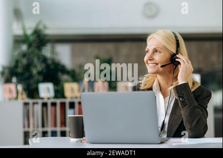 Elegante glückliche Geschäftsfrau arbeitet als Betreiber des Callcenters, sitzt am Schreibtisch im Büro. Reife weibliche Angestellte im Headset und stilvolle Anzug Beratung Kunden, lächelnd Stockfoto