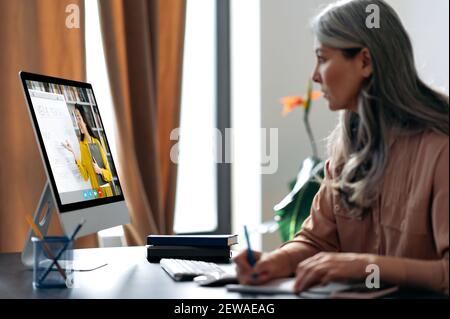 Fernunterricht. Konzentriert gealterte grauhaarige Frau, die online per Videoanruf studiert, nutzt App und Computer, sitzt an ihrem Schreibtisch, hört sich eine Online-Lektion an, auf dem Bildschirm ist der Lehrer in der Nähe Stockfoto