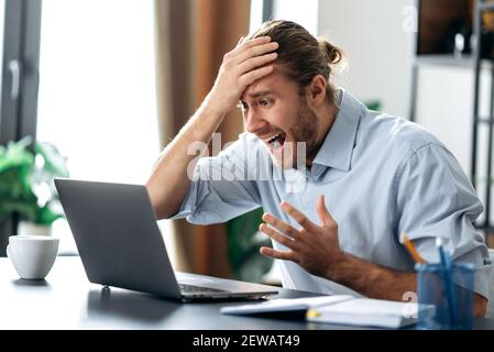 Schockiert verärgert kaukasischen beschäftigt Kerl Blick auf einen Laptop in Verwirrung, erhielt unerwartete Nachrichten, gescheitert einen Deal, schreit, hält seinen Kopf mit der Hand, sitzt am Schreibtisch Stockfoto