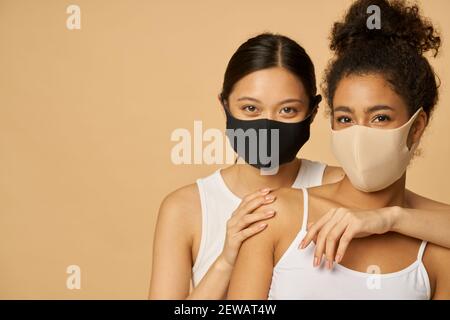 Porträt von zwei schönen jungen verschiedenen Frauen tragen schützende Gesichtsmasken Blick auf Kamera, während zusammen posiert isoliert auf beigem Hintergrund. Sicherheit, Pandemiekonzept Stockfoto