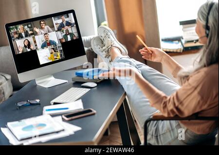 Ältere grauhaarige Frau sitzt zu Hause an einem Tisch mit ihren Beinen auf den Tisch geworfen, hört sich eine Online-Lektion per Videokonferenz mit Computer, Lehrer und Schüler sind auf dem Bildschirm, Abstand Stockfoto