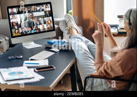 Telearbeit von zu Hause aus. Moderne ältere grauhaarige Frau mit Kollegen online per Videoanruf kommunizieren nutzt App und Computer, sitzen in ihrem Home-Office, auf dem Bildschirm viele verschiedene Menschen Stockfoto