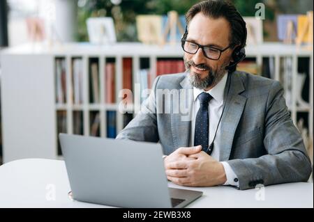 Glücklicher kaukasischer Geschäftsmann bei einer Videokonferenz, im Gespräch mit Kollegen über den Arbeitsprozess. Stilvoller, reifer Manager in Headset und Brille sitzt mit einem Laptop am Schreibtisch und lächelt Stockfoto