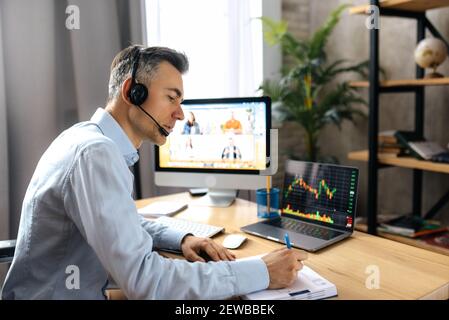 Online-Lektion oder Business-Webinar. Erfolgreicher Mann sitzt am Schreibtisch macht Notate während Online-Meeting, auf einem Computer-Bildschirm sind verschiedene multirassische Menschen in einem Online-Vortrag von gesammelt Stockfoto